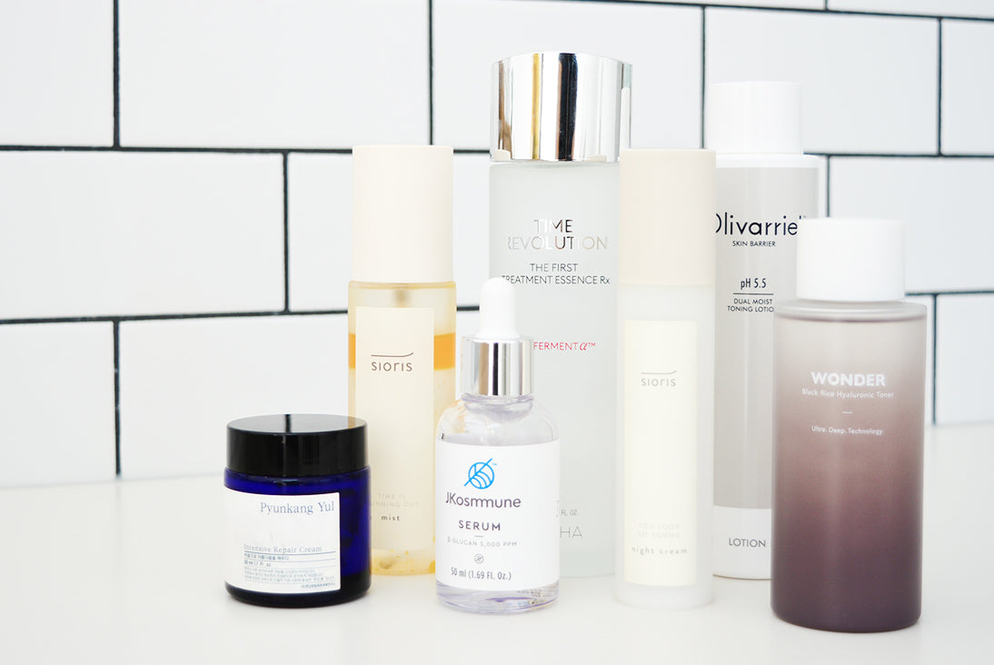 A collection of seven different skincare products in bottles and jars of various sizes and colors, arranged together against a white subway tile background.