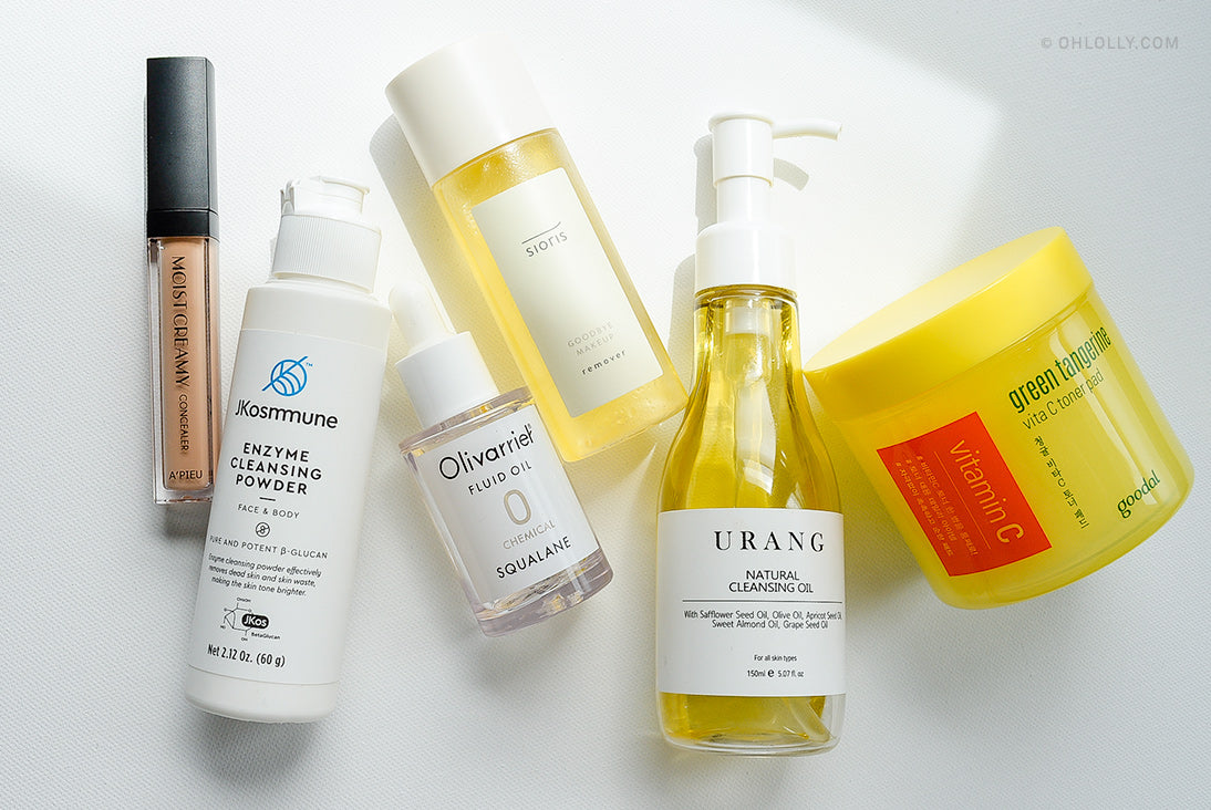 An overhead flat lay of various skincare and makeup products, including bottles, a jar, a dropper, and a concealer tube, arranged on a white surface with soft shadows.