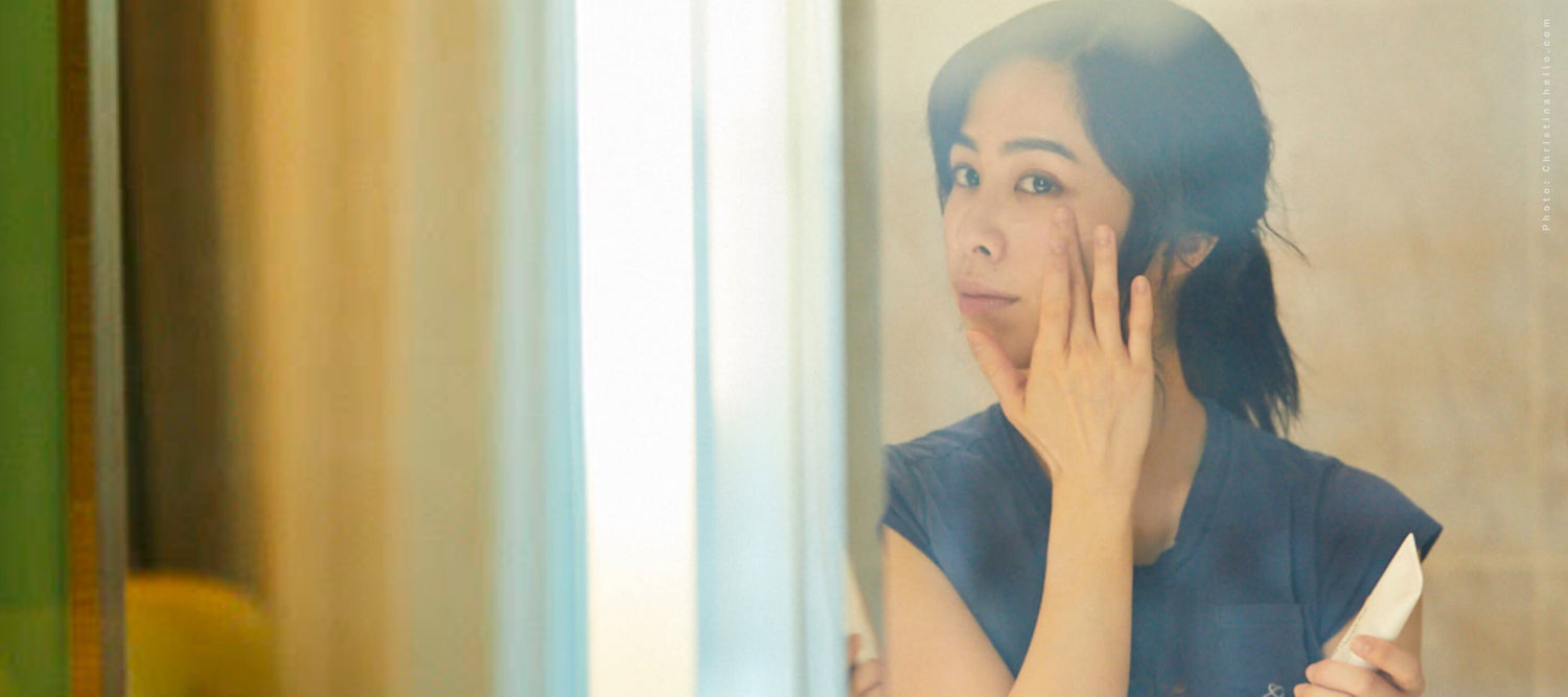  A woman with dark hair pulled back looks into a mirror while applying skincare to her cheek in a sunlit room.