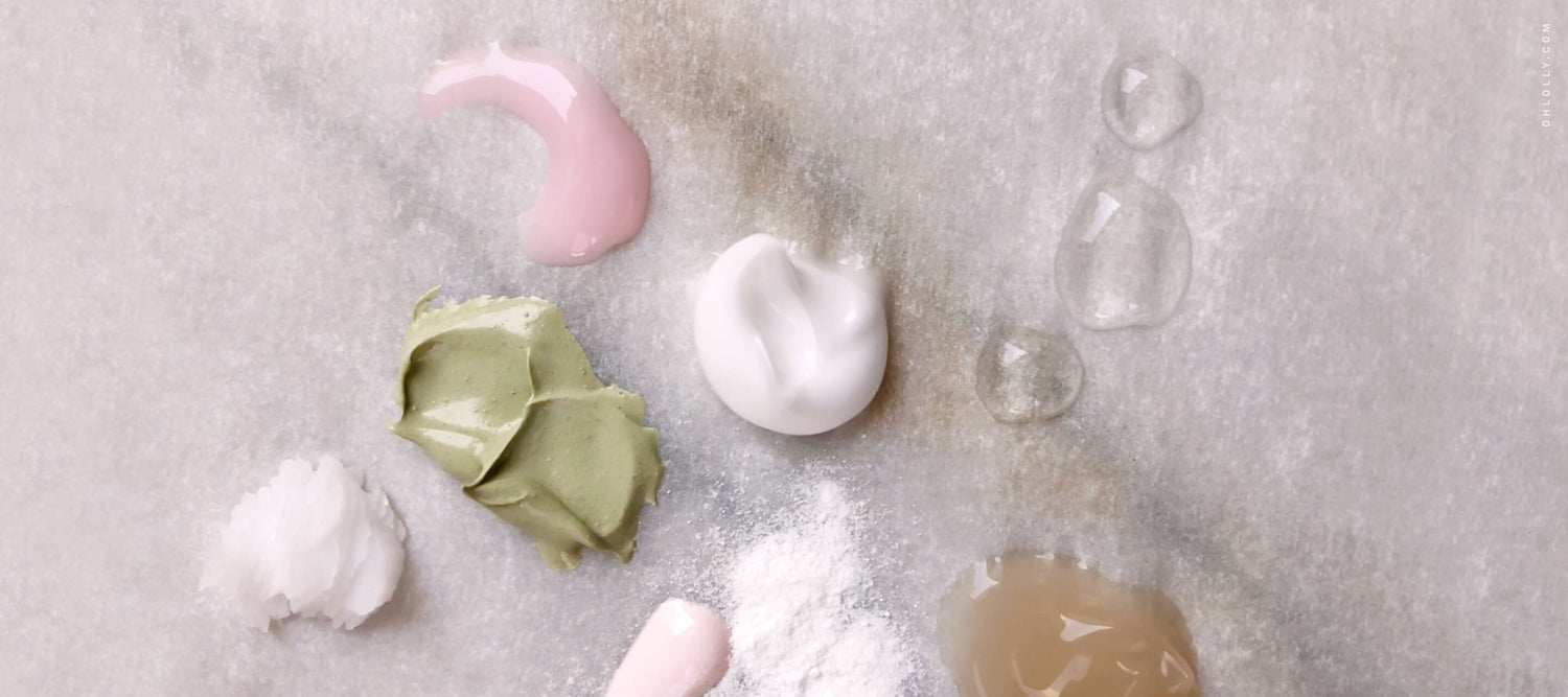  Various dollops and smears of different skincare product textures, including green, pink, and white creams and clear gels, on a marble surface.