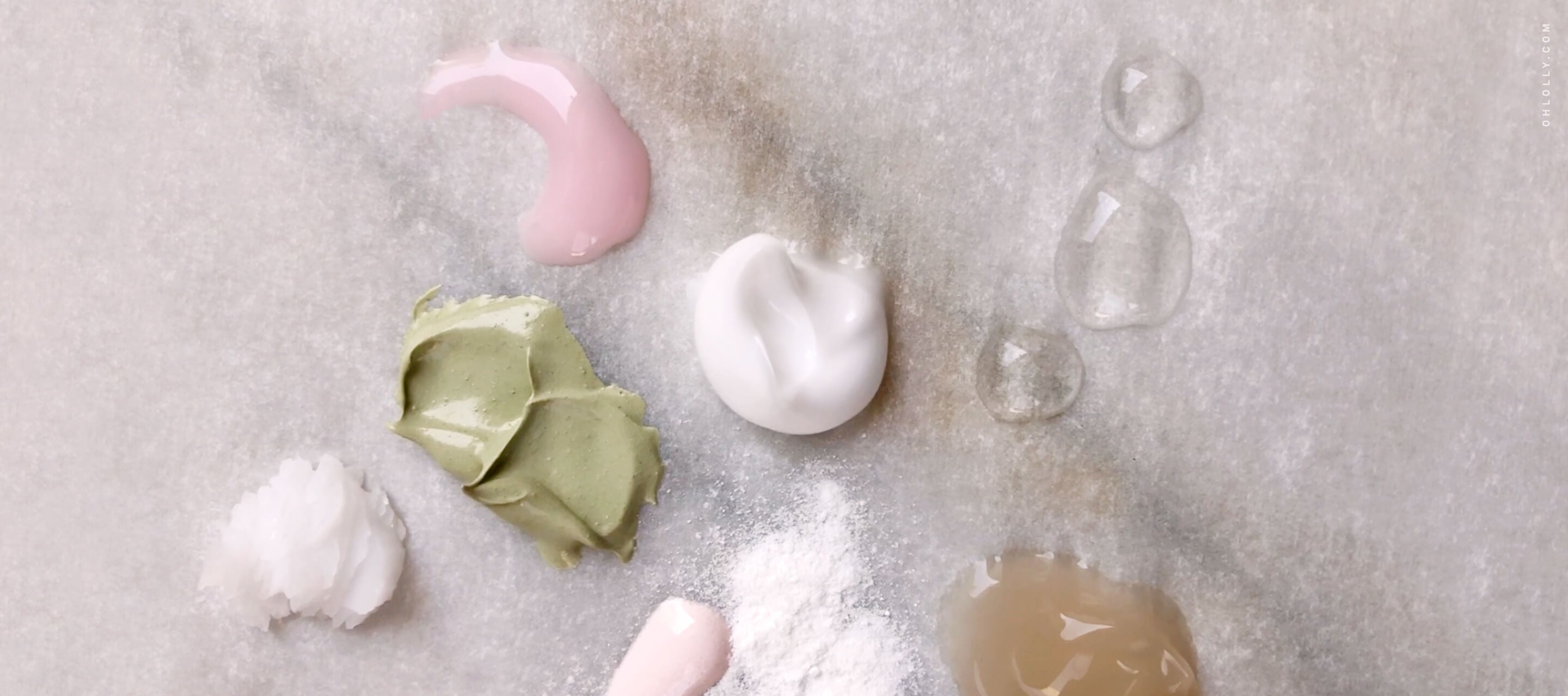  Various dollops and smears of different skincare product textures, including green, pink, and white creams and clear gels, on a marble surface.