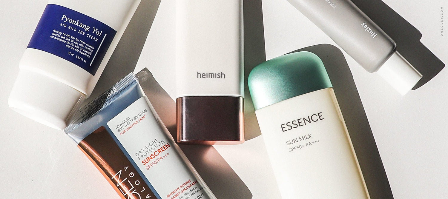 A flat lay arrangement of five different tubes of sunscreen, including brands like Pyunkang Yul, Neogen, and Missha, angled across a white surface with shadows.