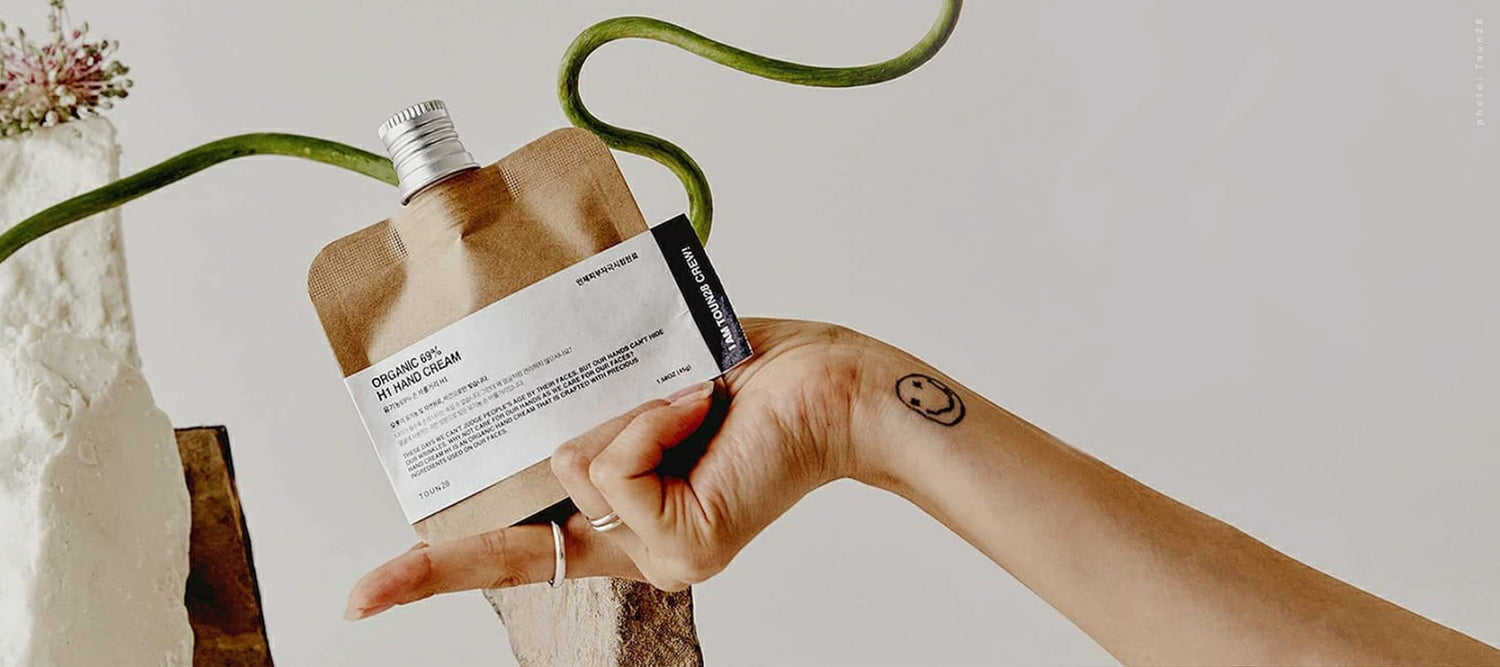  A person's hand holding a Toun28 sustainable skincare pouch against a white background with a green vine, featuring a small smiley face tattoo on the wrist.