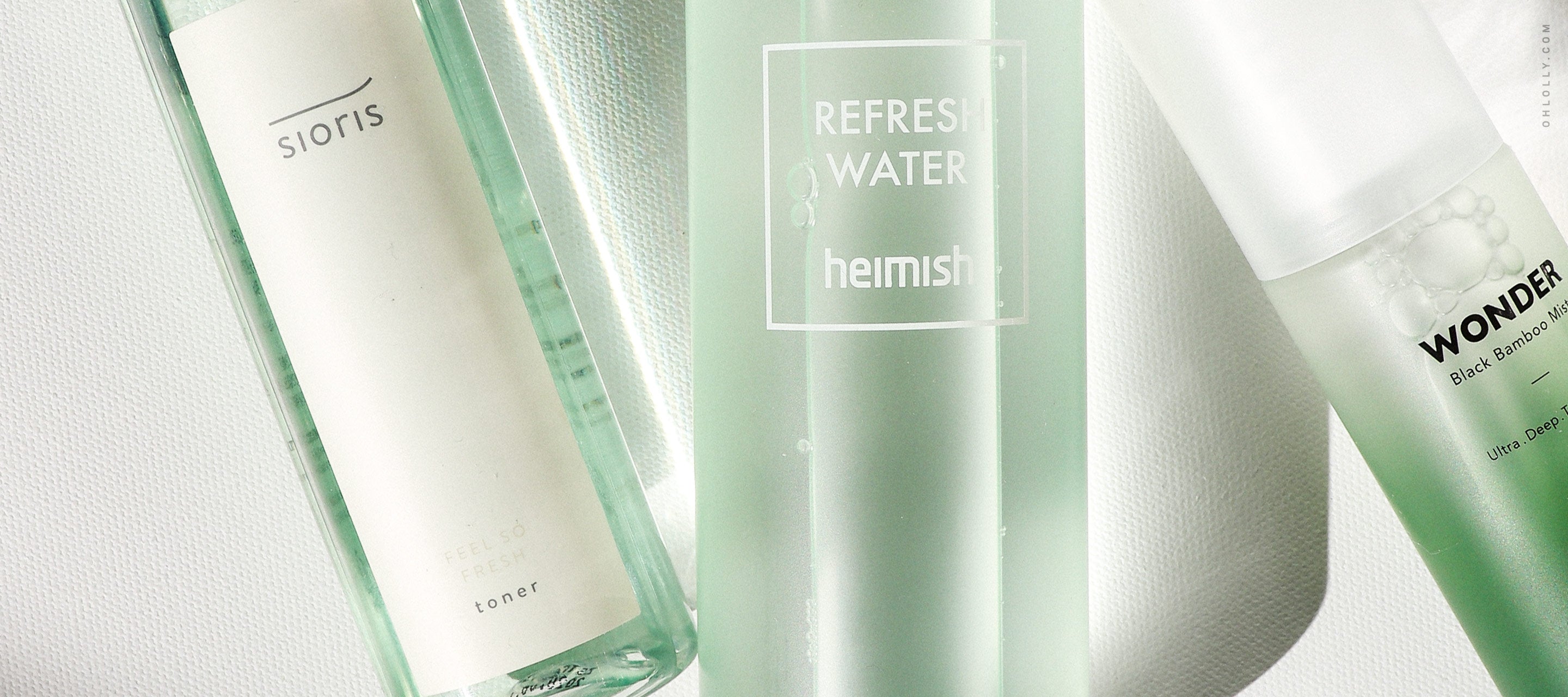 A close-up of three green-tinted skincare bottles from brands including Sioris, Heimish, and Haruharu Wonder, arranged horizontally on a white surface.