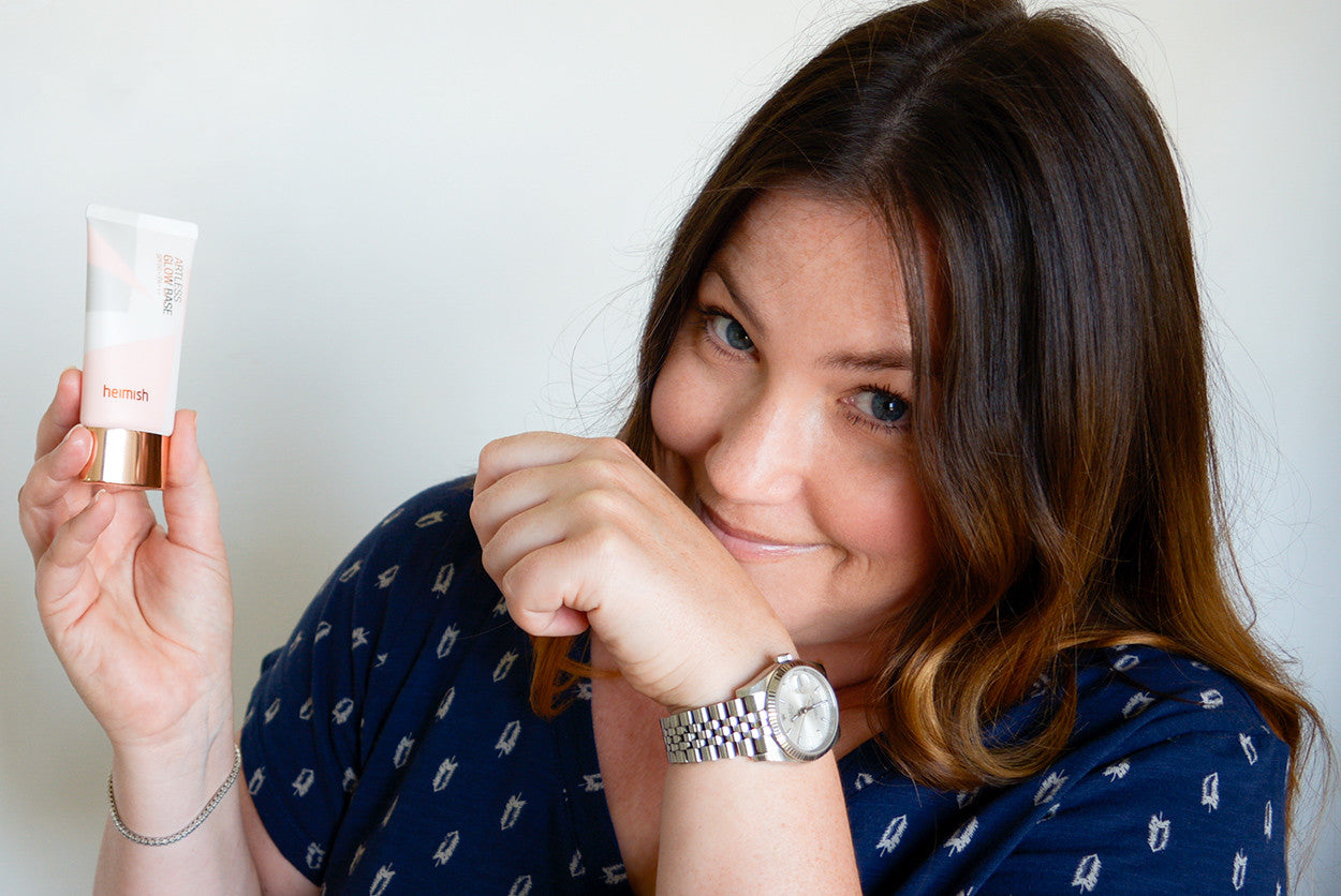 A woman holds a tube of Heimish Glow Base and smiles at the camera, wearing a watch.