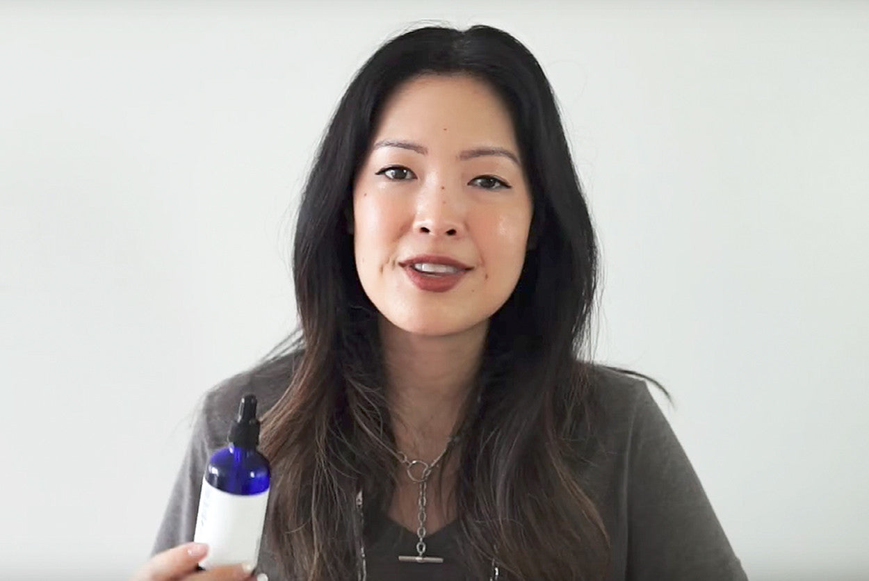A woman with dark hair holds a blue glass dropper bottle and looks at the camera.