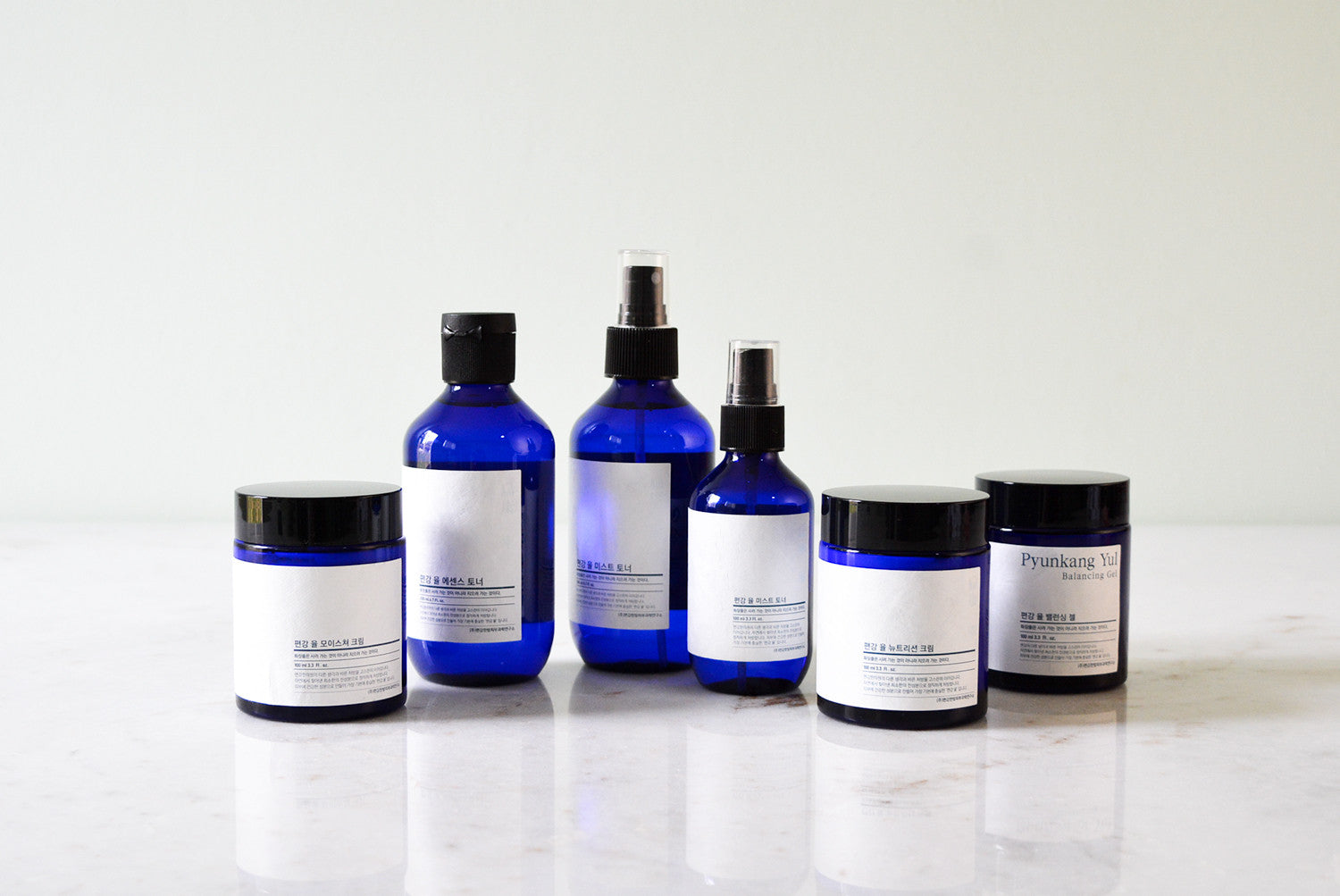 A lineup of six Pyunkang Yul skincare products in blue glass containers with white labels, including jars, spray bottles, and dropper bottles, against a white background.