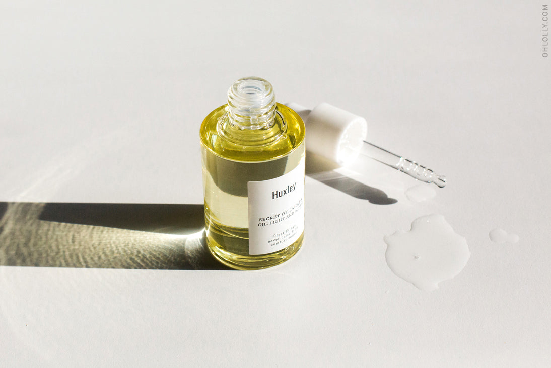  An open glass bottle of yellow Huxley body oil next to its dropper, with a small spill on a white surface in bright sunlight.