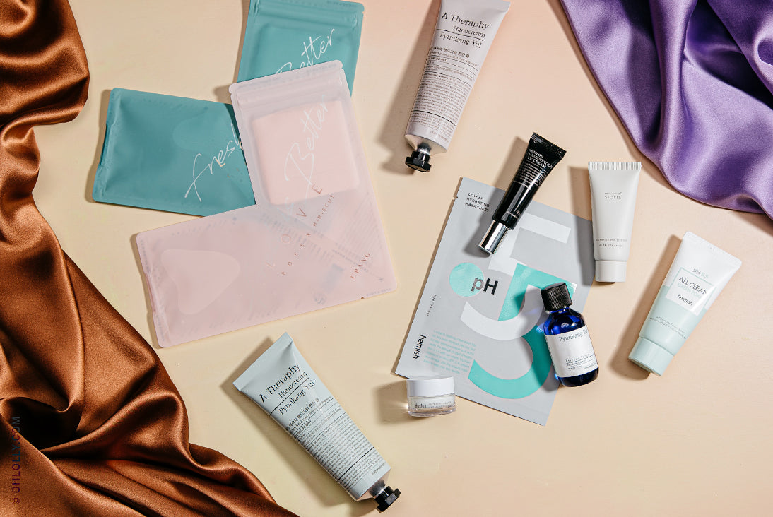 An assortment of various skincare products, including tubes, small bottles, and colorful pouches, flat-laid on a tan surface with draped brown and purple satin fabric.