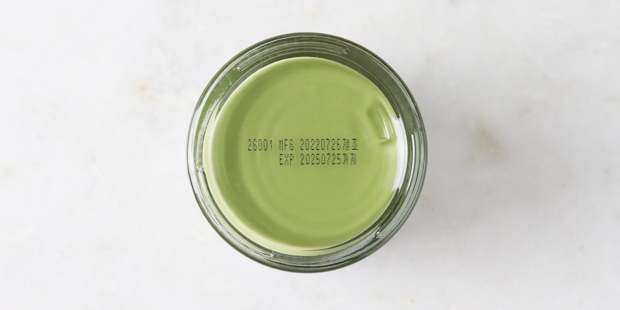  Top-down view of a round glass jar filled with a thick green cream, showing a batch code and expiration date printed on the bottom of the container.