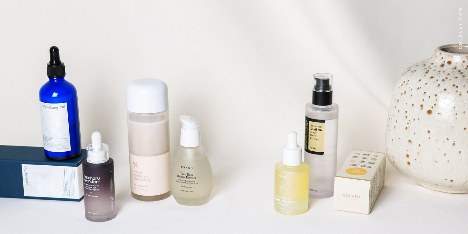  A collection of six K-beauty skincare products, including various serums and toners in bottles, arranged on a white surface next to a speckled ceramic vase.