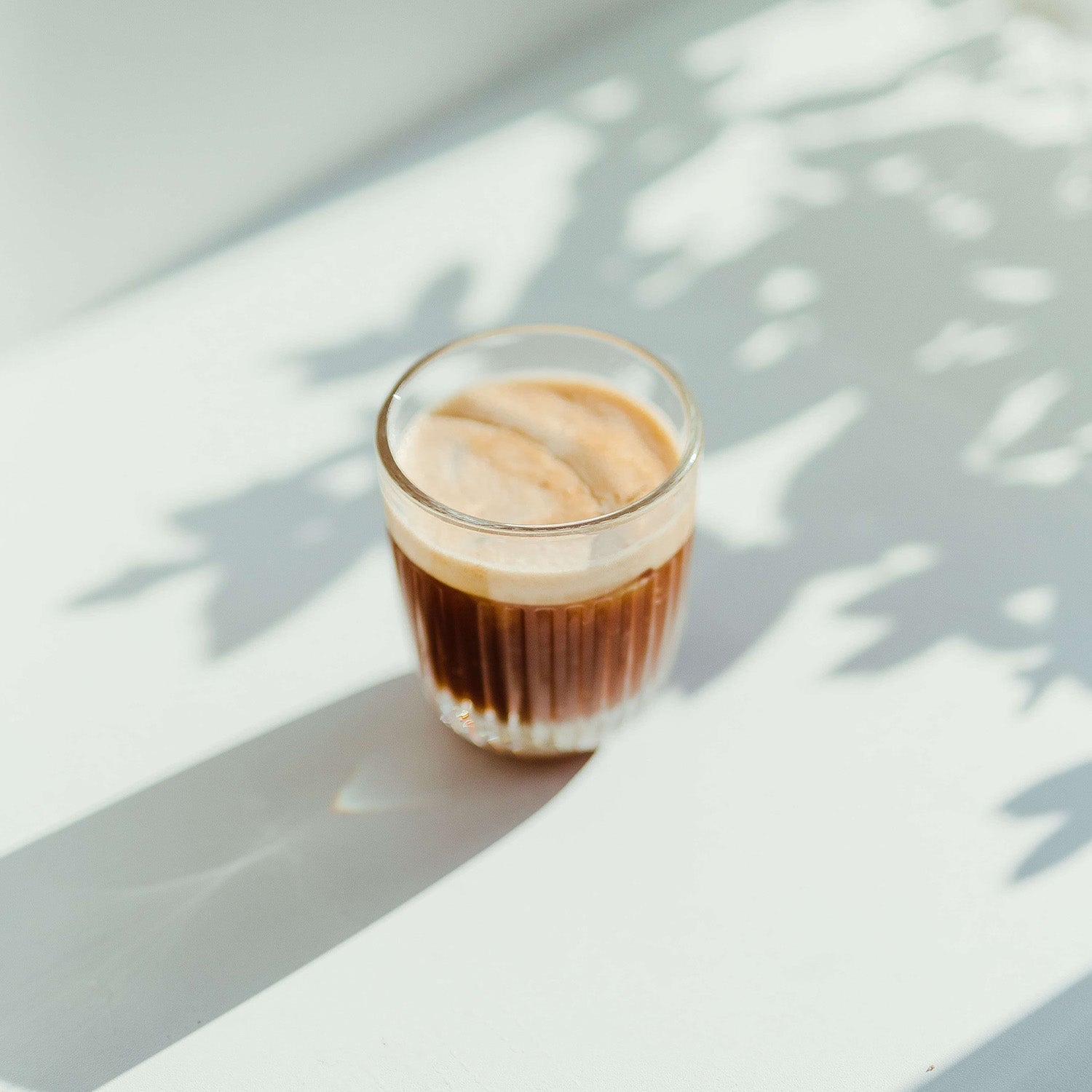  A small glass of espresso with a layer of crema on top, sitting on a white surface with leafy shadows from sunlight.