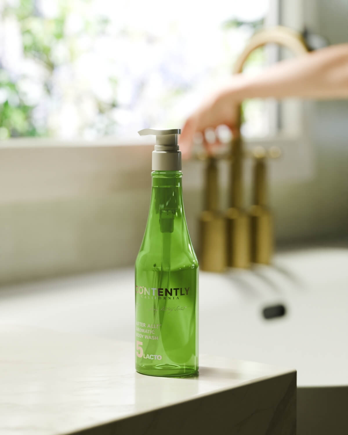 A green bottle of CONTENTLY CALIFORNIA's "After All! Aromatic Body Wash" is placed on the edge of a white kitchen sink with a blurred golden faucet and window in the background.