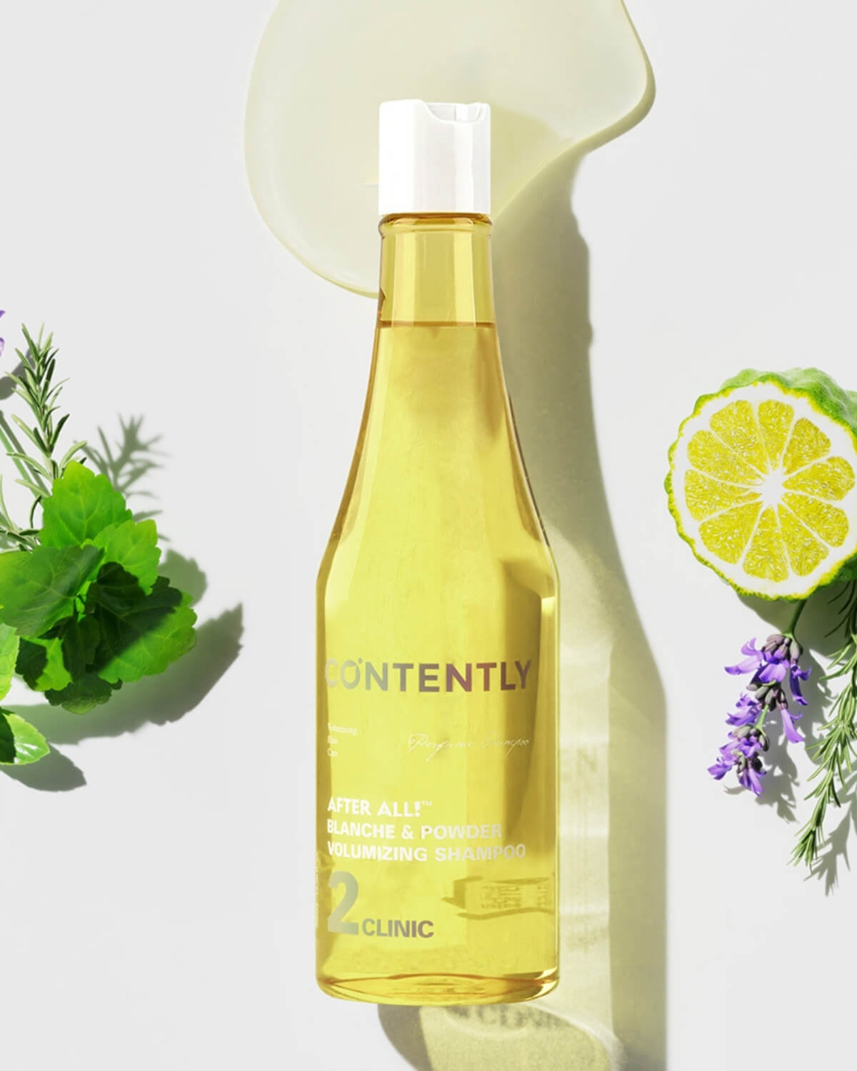 The yellow shampoo bottle is centered on a white background with a smear of the clear yellow product behind its cap. It is flanked by a halved bergamot citrus slice and lavender on the right, and green herbal sprigs on the left.