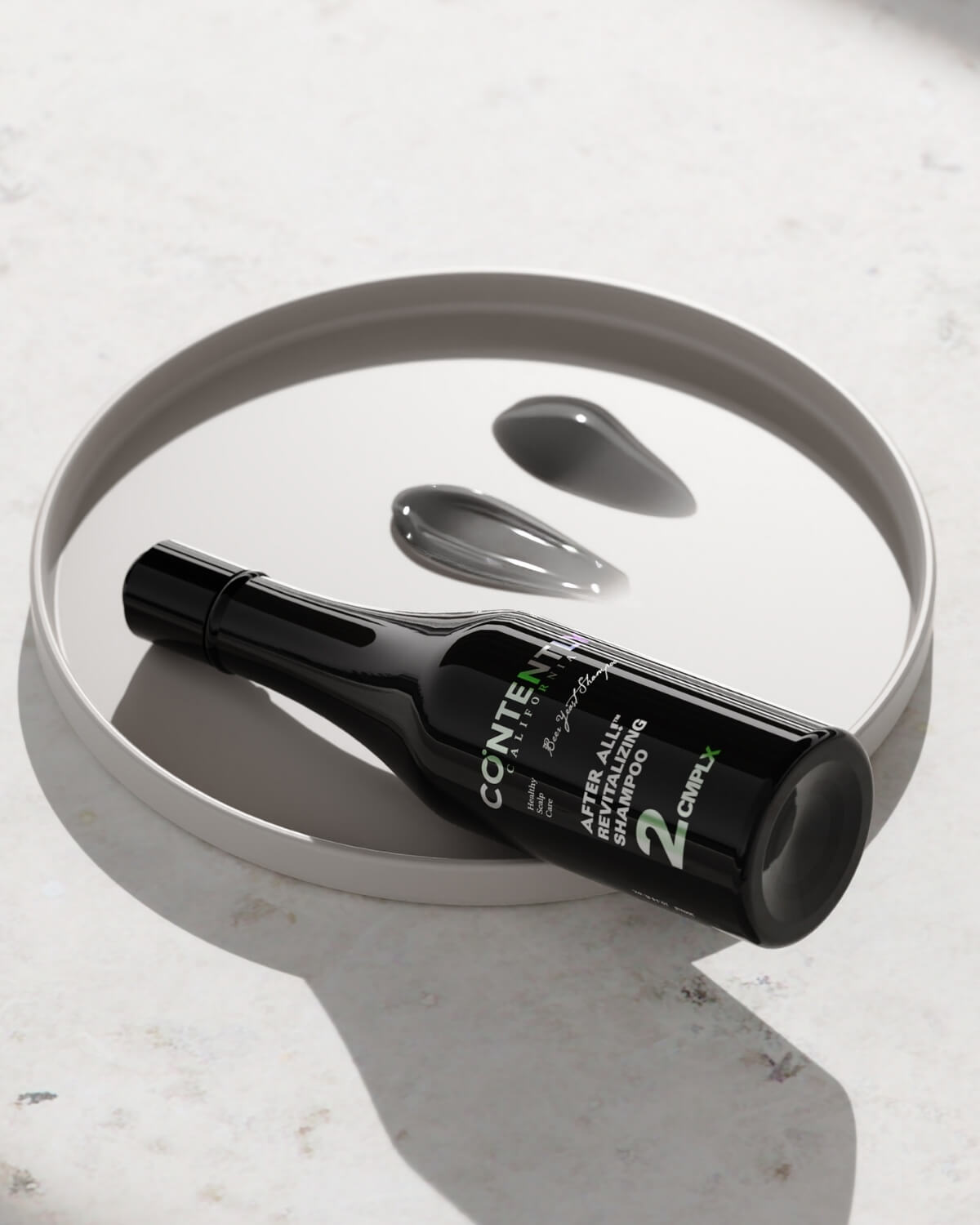 The black shampoo bottle is resting on a white, round tray next to two glossy dark gray/black swatches of the shampoo product.