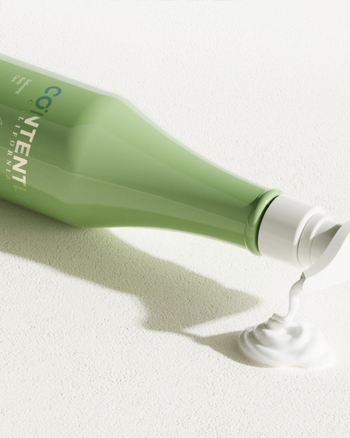 A close-up, horizontal shot of the light green CONTENTLY CALIFORNIA body lotion bottle. The cap is open, and a stream of white, creamy lotion is being dispensed onto a textured white surface, forming a dollop of product.
