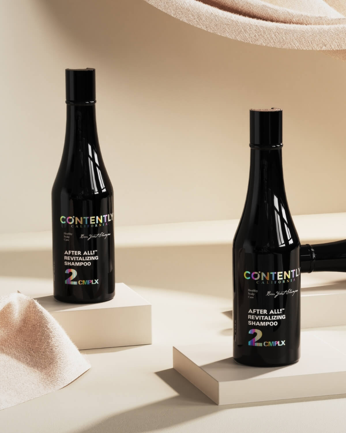 Two bottles of the Contently Revitalizing Shampoo are displayed on white pedestals in a warm, light-beige setting with draped fabric.