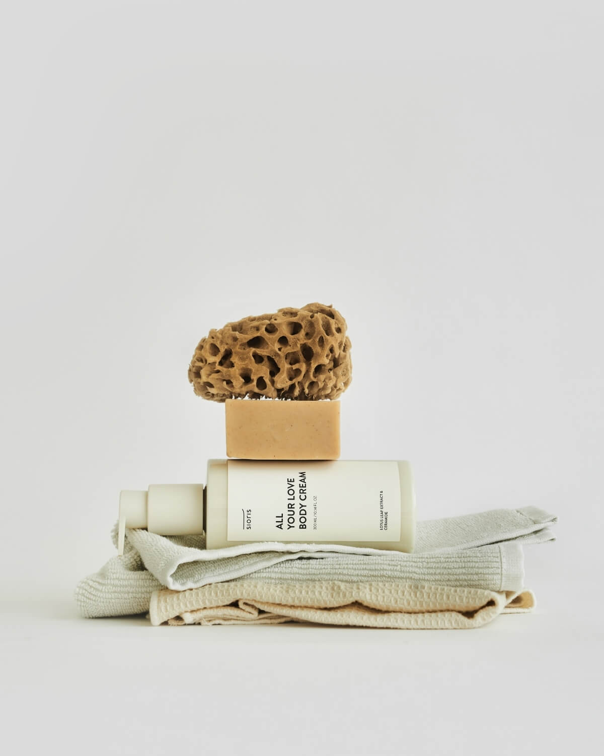 Sioris All Your Love Body Cream bottle layered horizontally between folded bath towels, soap, and sponge.