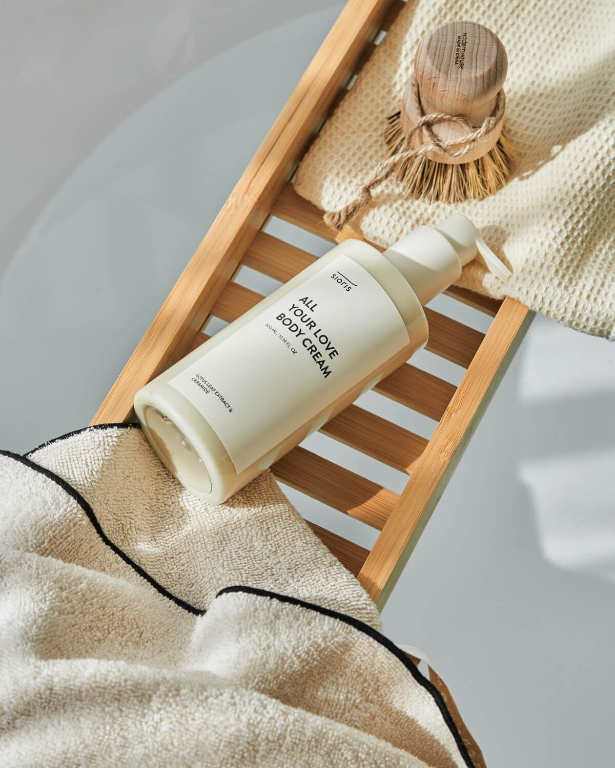 Sioris All Your Love Body Cream bottle on a bamboo bathtub caddy tray alongside a wooden bath brush.