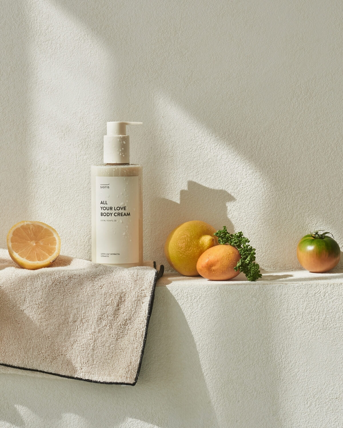 Sioris All Your Love Body Cream bottle on a sunny ledge with fresh citrus fruits, a tomato, and a towel.