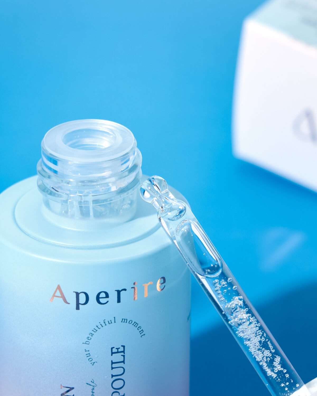 A macro close-up of the ampoule bottle's neck, showing the dropper filled with a clear, bubbly serum and resting against the glass opening. The background is a bright blue.
