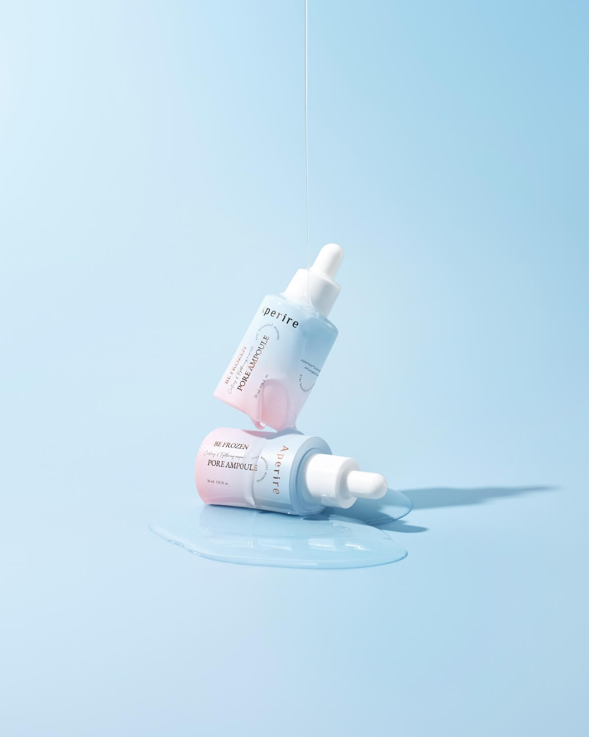 Two ombre ampoule bottles are creatively arranged on a light blue surface. One bottle is suspended above the other lying on its side in a pool of clear liquid, with a thin string of liquid dripping from above.