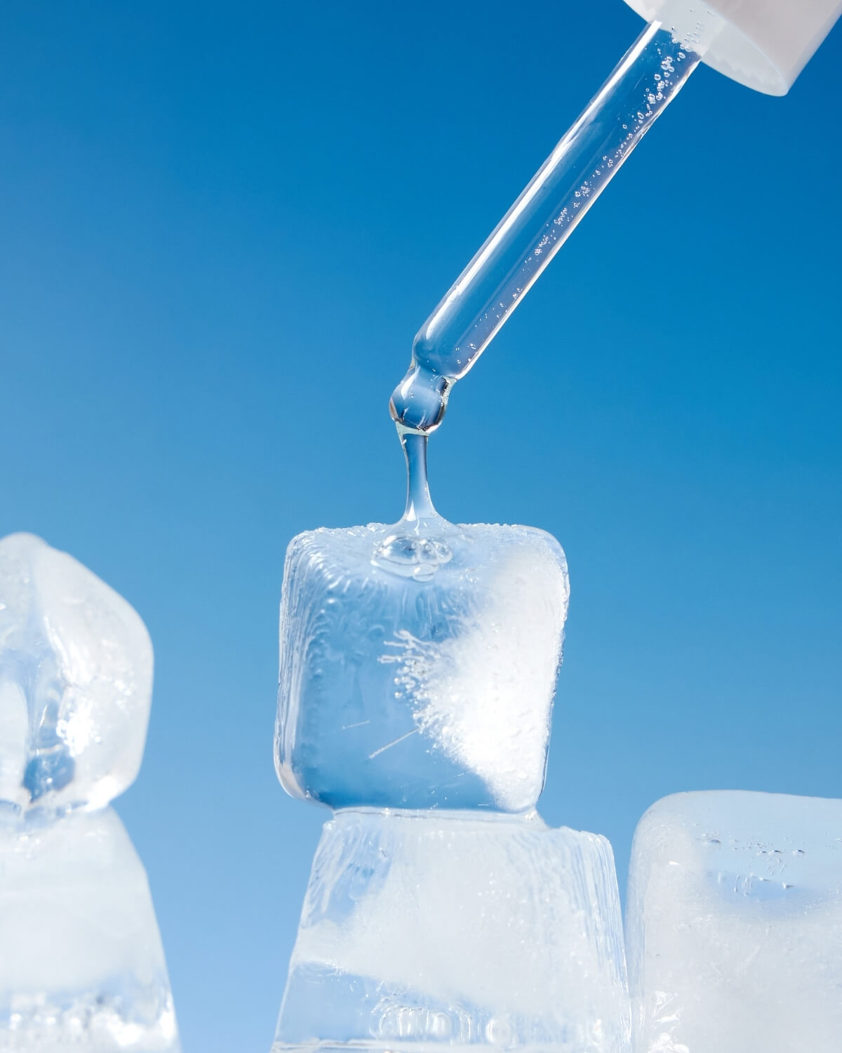  A close-up, dramatic shot of a glass dropper dispensing a clear liquid onto a stack of three ice cubes against a vibrant blue background, emphasizing the "Be Frozen" cooling concept.