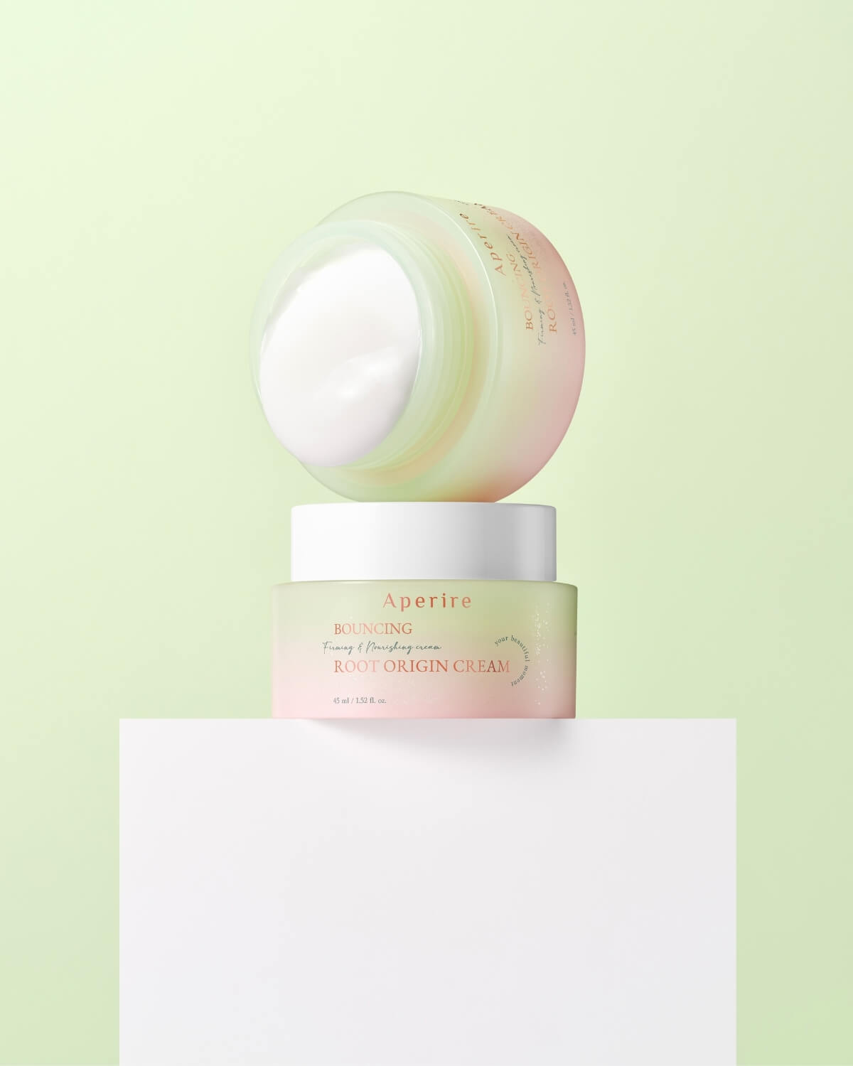 Two jars of Aperire Bouncing Root Origin Cream are stacked on a white platform against a pale green background. The bottom jar is closed, while the top jar is opened, revealing the thick, white cream inside.