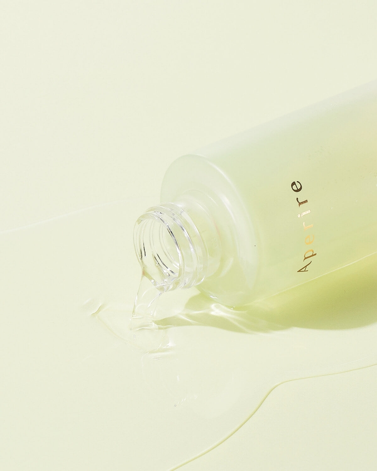 An artistic display of the toner bottle and a single cotton pad on a textured, rough white surface. A small, clear drop of the amber liquid is falling onto the cotton pad.