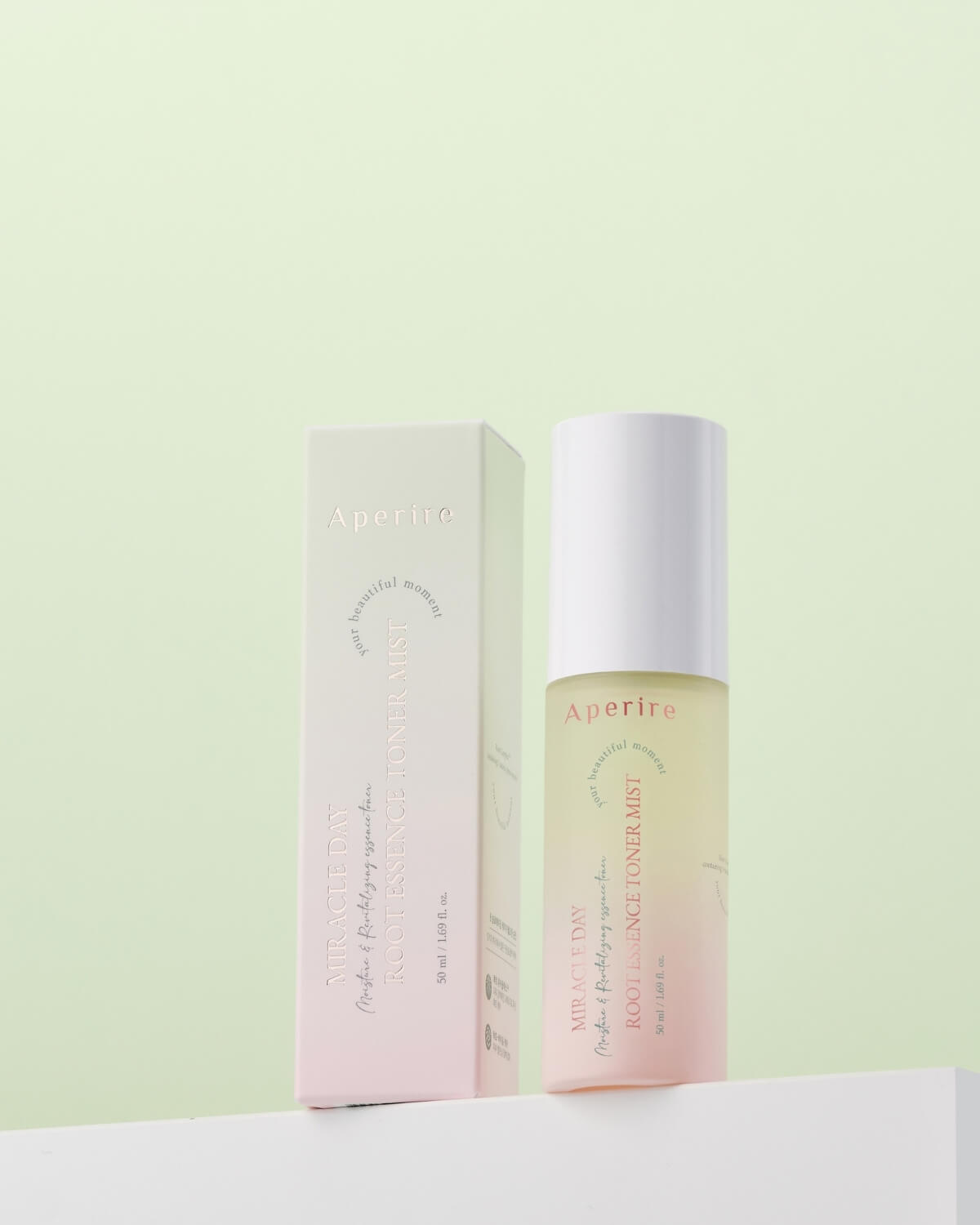 A bottle of Aperire Miracle Day Root Essence Toner Mist next to its pink and white box, set against a light green background. The product is a bi-phase mist with a pale yellow layer on top and a pink layer on the bottom.