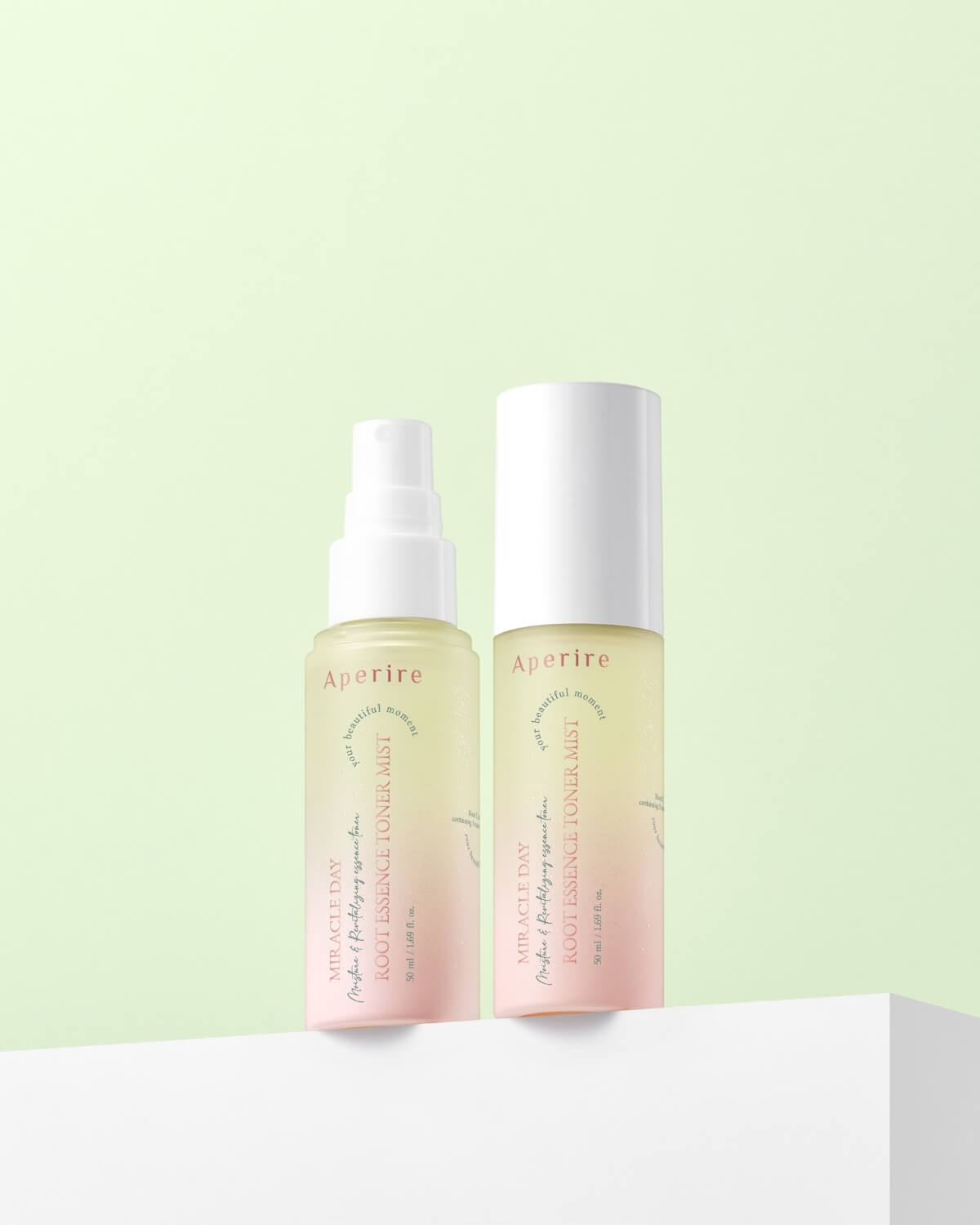 Two bottles of Aperire Miracle Day Root Essence Toner Mist on a white platform against a light green background. One bottle has a white spray pump, and the other has a solid white cap. Both show the bi-phase yellow and pink liquid.