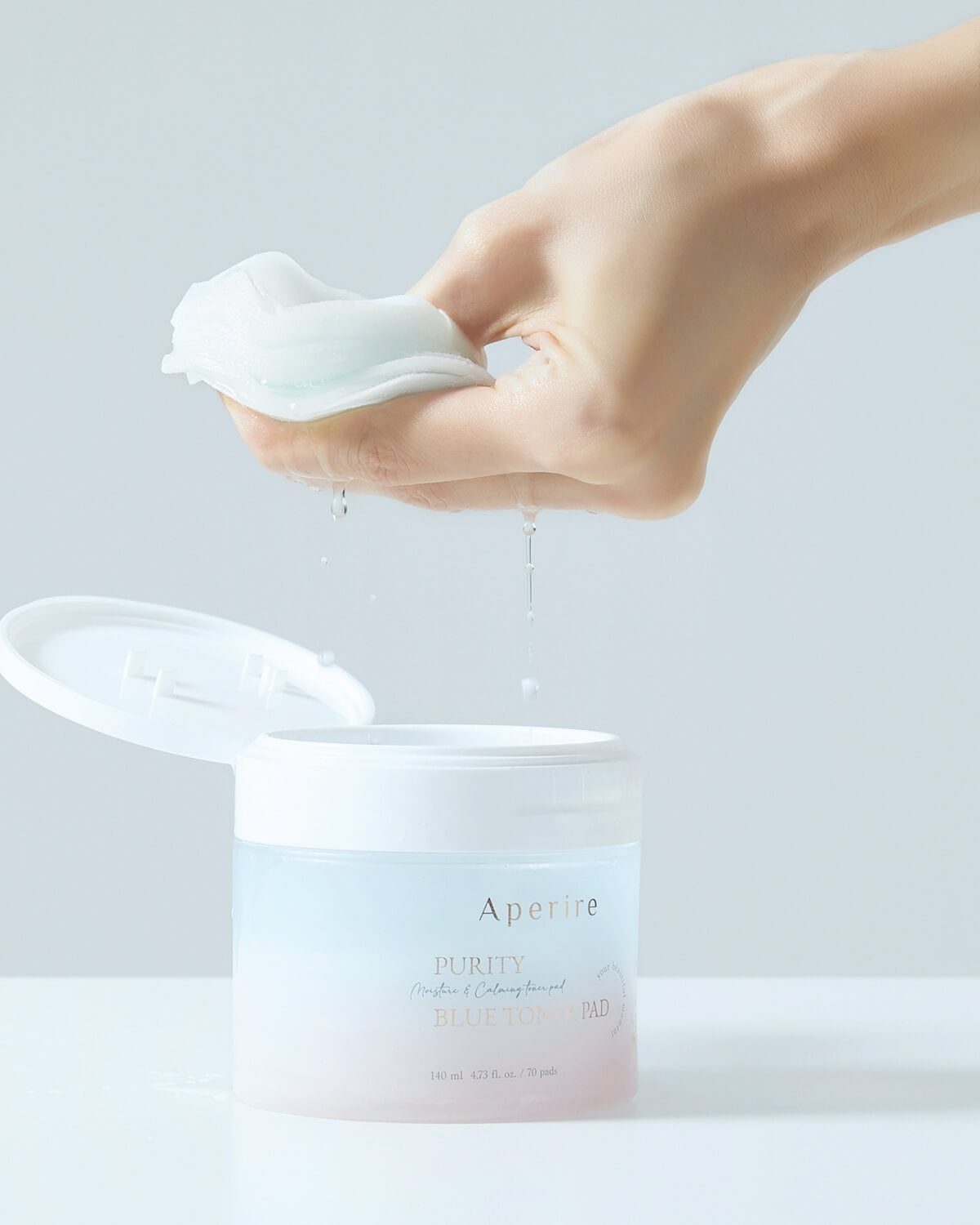 A hand wrings out a stack of three saturated toner pads above the open container. Drops of liquid fall back into the jar, demonstrating the toner's abundant moisture.