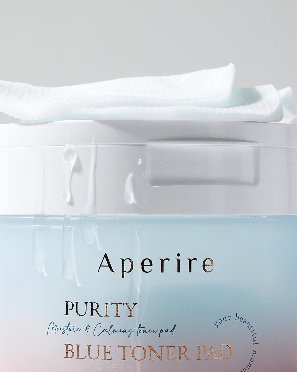 A macro close-up of the Aperire Purity Blue Toner Pad container's lid with a few pads resting on top. Drops of liquid are visible, emphasizing the moisture.