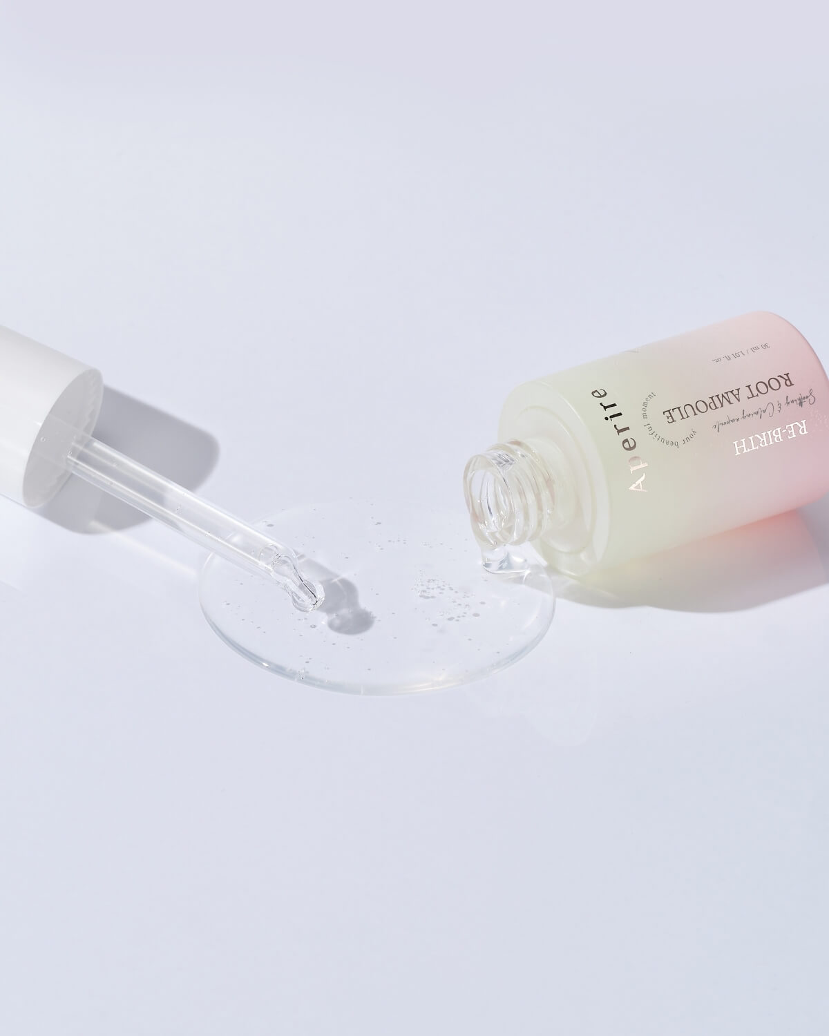 The bottle of Aperire Re-birth Root Ampoule is lying on its side on a white surface, with a small pool of the clear serum visible. The glass dropper is extended next to it, showing the product's translucent, watery texture.