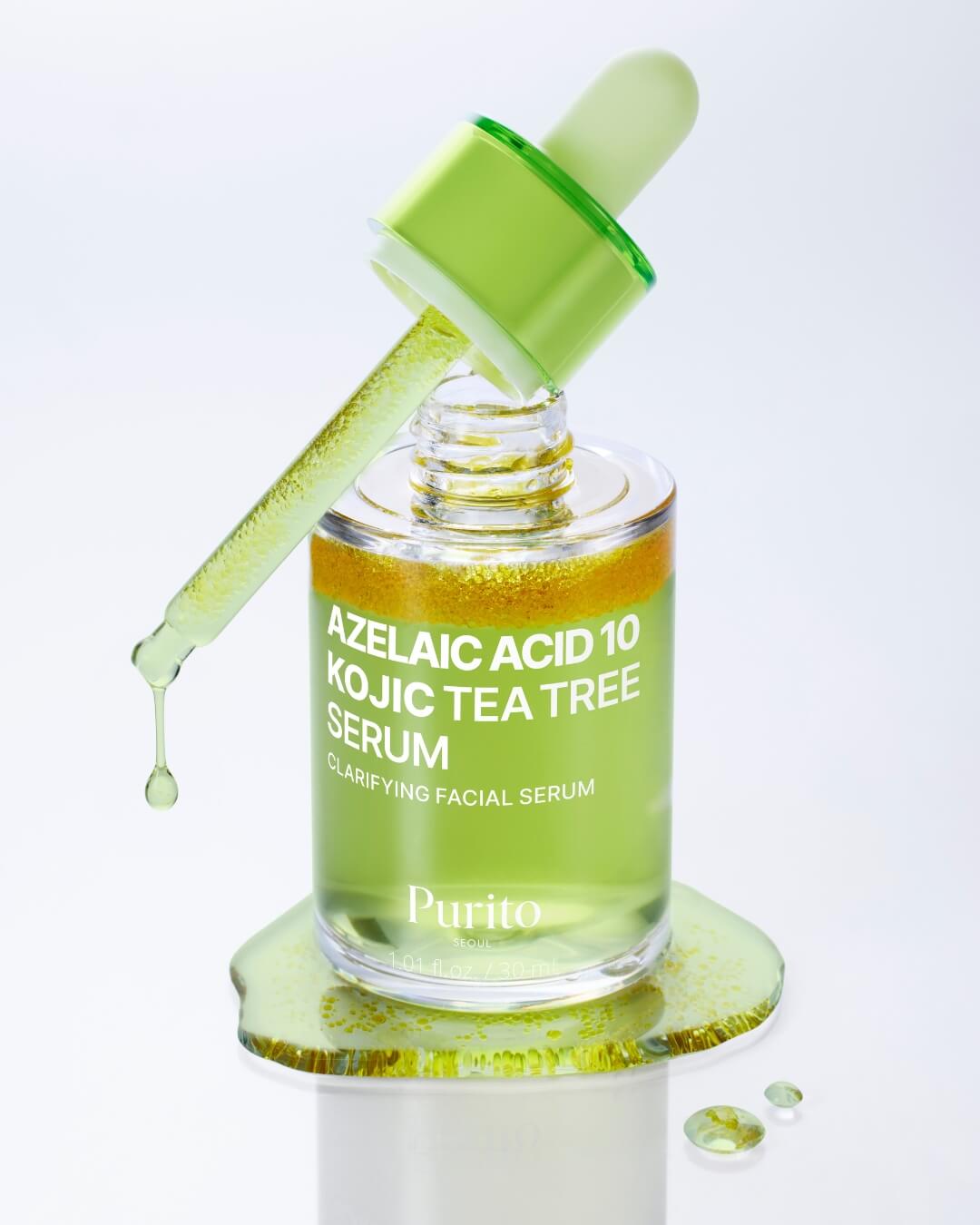 An angled studio shot of the Purito Azelaic Acid 10 Kojic Tea Tree Serum. The bottle sits in a puddle of the green serum, and the dropper is held out, dripping a single drop of the bi-phase liquid.