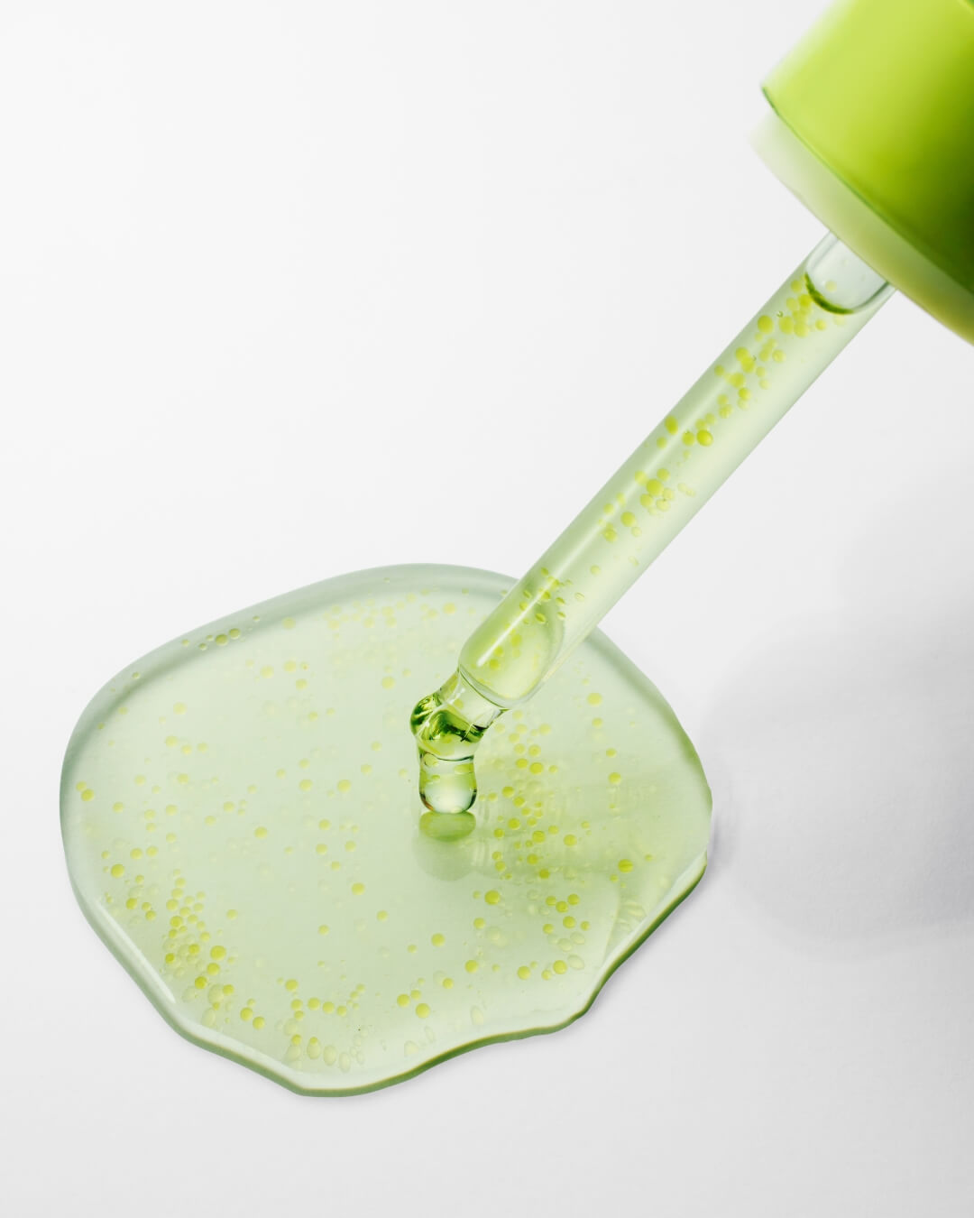A close-up detail shot of the serum dripping onto a white surface. The green-yellow liquid is spread out, clearly showing the suspended, lighter yellow oil droplets within the serum. The dropper from the bottle is touching the center of the puddle.