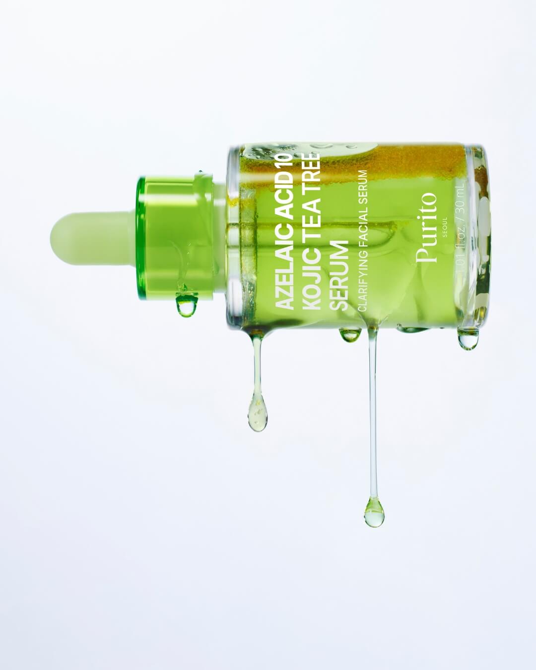 A close-up studio shot of the Purito Azelaic Acid 10 Kojic Tea Tree Serum bottle lying on its side. Large droplets of the green serum are hanging and dripping from the bottle, showcasing the liquid's clarity and color.