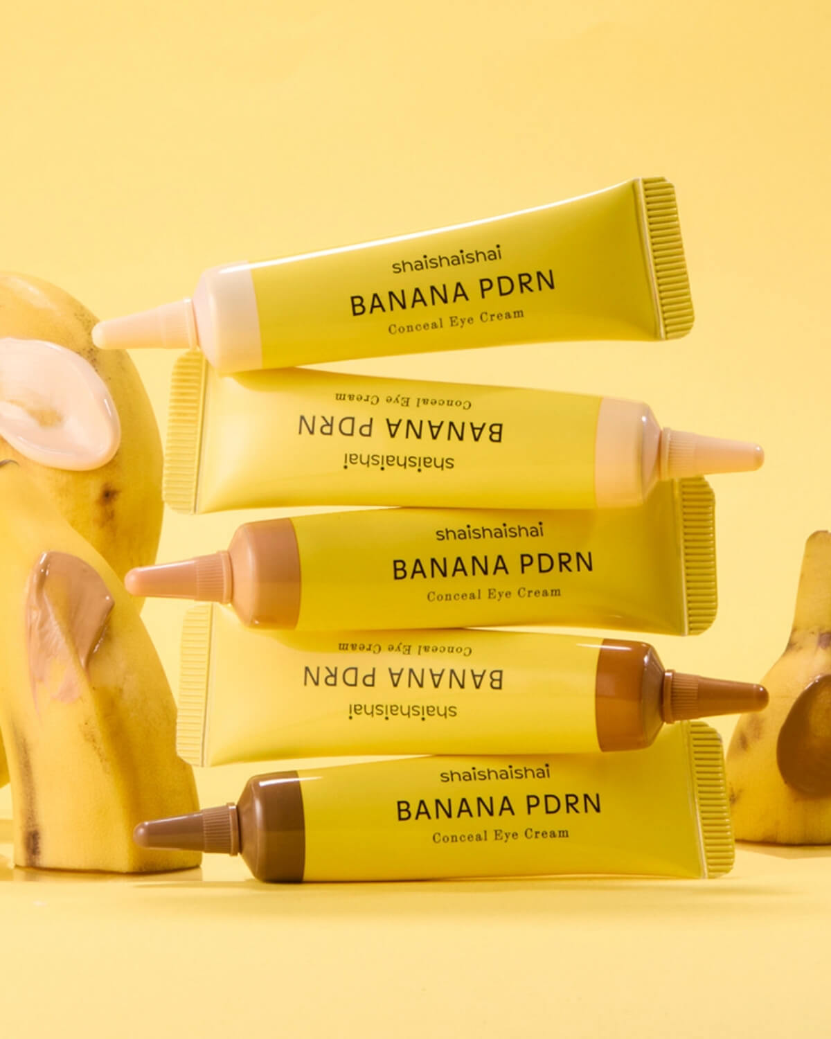 Stacked tubes of Banana PDRN Conceal Eye Cream balanced against fresh banana slices on a yellow backdrop.