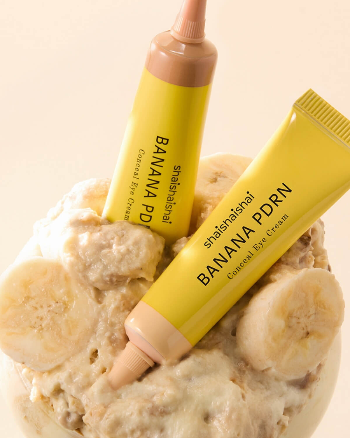 Two tubes of shaishaishai Banana PDRN Conceal Eye Cream tucked into a bowl of mashed fresh bananas.