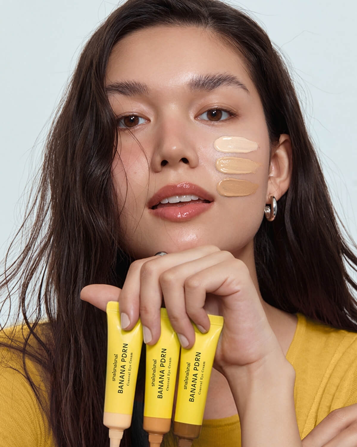 A model with three shades of Banana PDRN Conceal Eye Cream swatched on her cheek for color matching.