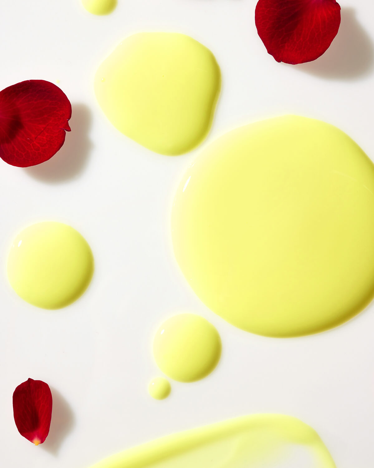 Close-up of yellow serum texture droplets and red rose petals on a clean white surface.
