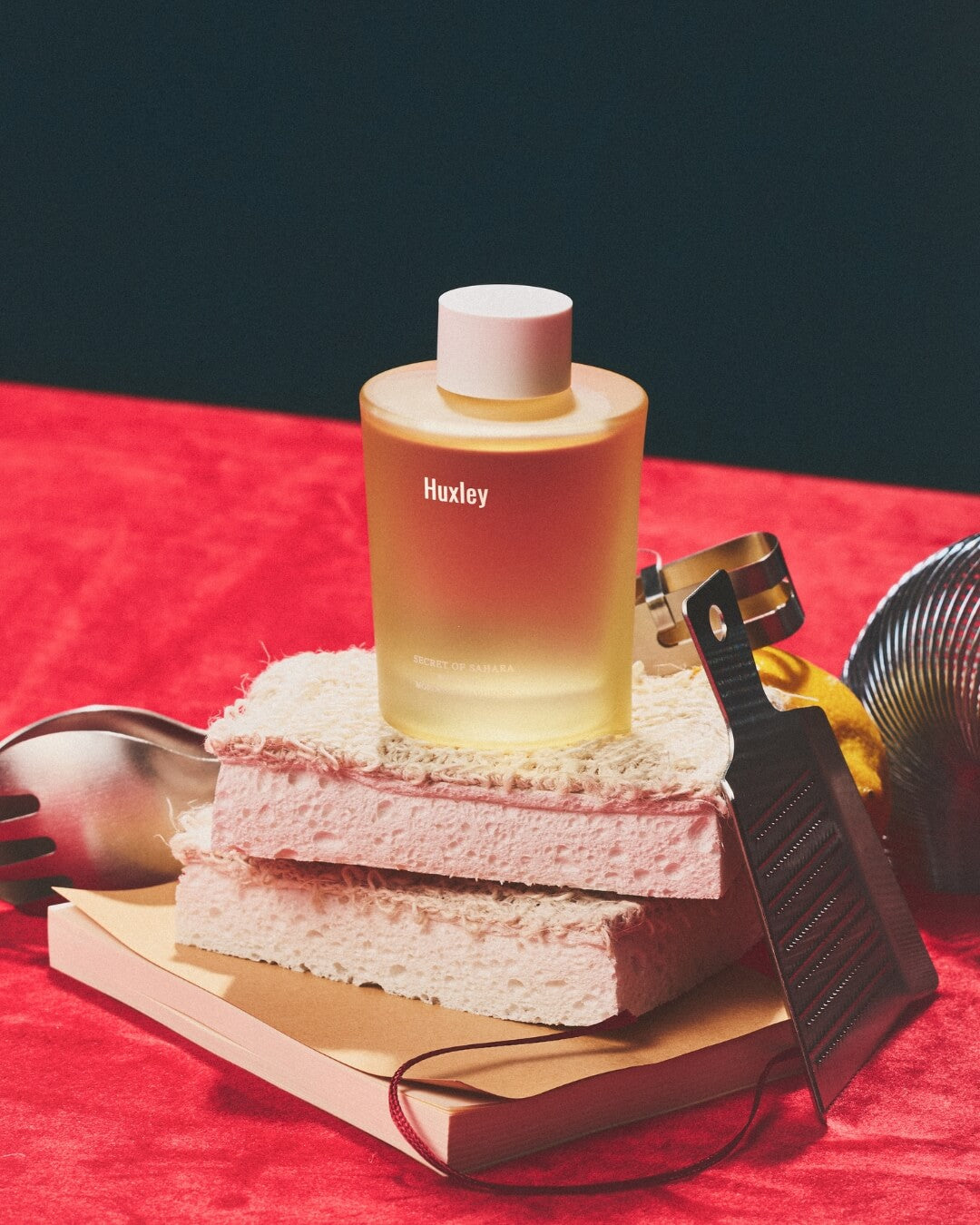 Huxley Moroccan Gardener body oil sitting on stacked pink bath sponges over a red surface.