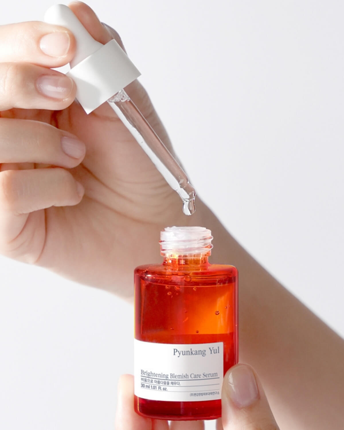 A close-up of hands holding the Pyunkang Yul Brightening Blemish Care Serum bottle, with the dropper being pulled out and a drop of the orange-red serum suspended at the tip.