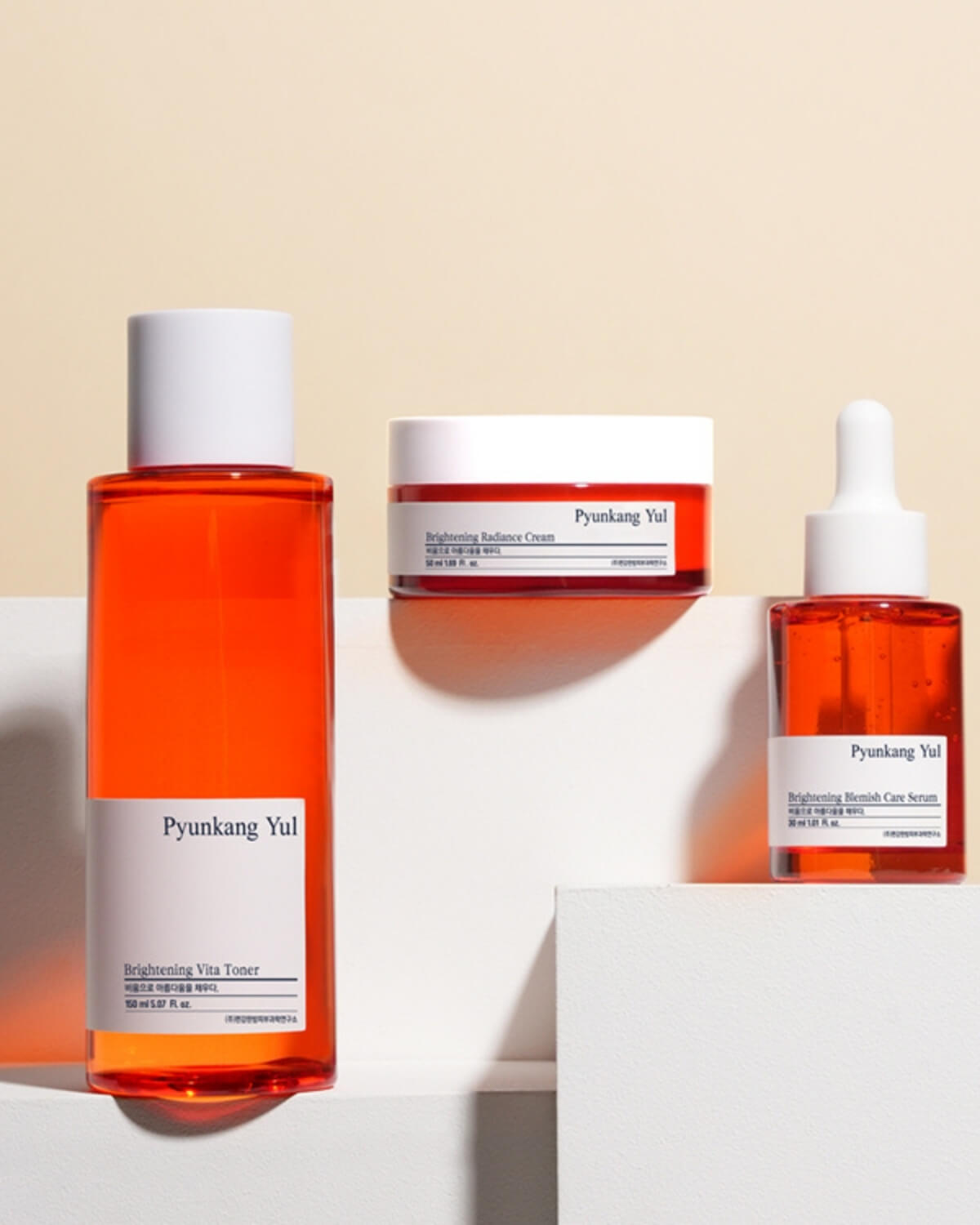 The three Pyunkang Yul Brightening products (Vita Toner, Radiance Cream, and Blemish Care Serum) arranged on white geometric blocks against a warm beige background.