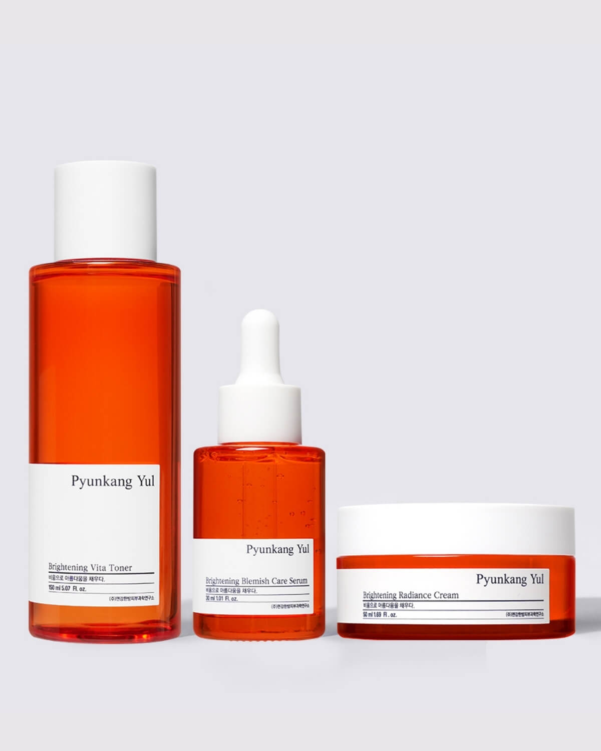 A clear, full-product shot of the three-piece Pyunkang Yul Brightening skincare line: the tall Vita Toner, the Blemish Care Serum with a dropper, and the Radiance Cream jar. All have an orange-red tint.