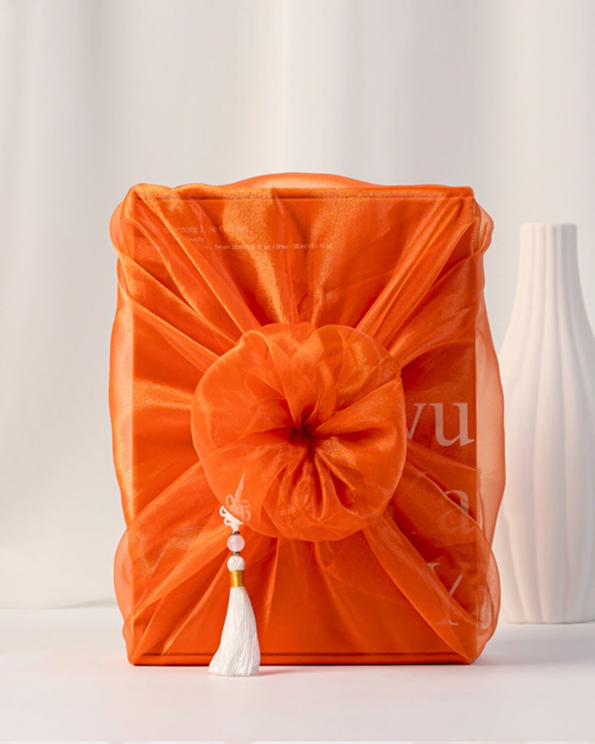 A gift box or product set wrapped in a vibrant, sheer orange bojagi with a large, ruffled knot in the center and a white tassel charm hanging from it. The box sits on a white surface next to a white vase.