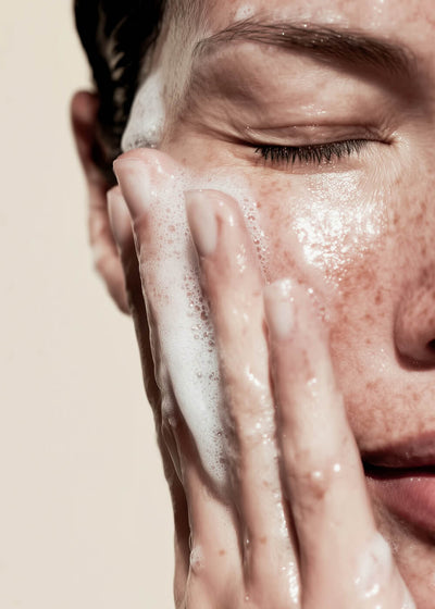 Person washing their face with a soapy hand against a beige background