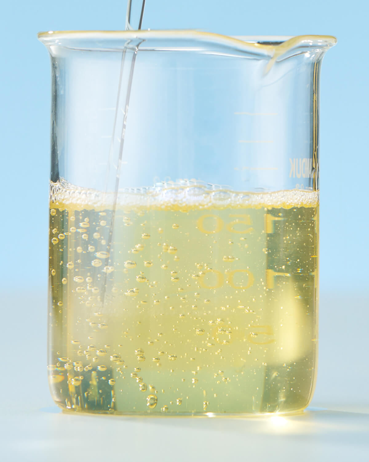 A close-up shot of the yellow liquid cleanser being poured or mixed inside a clear lab beaker, showing small, light bubbles on the surface.