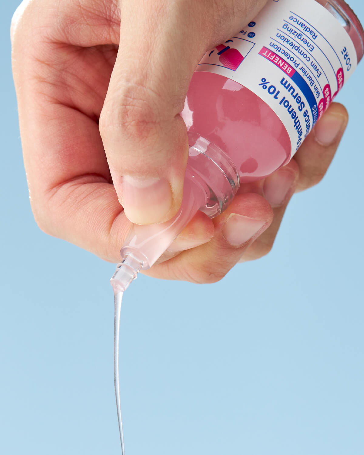 A hand gently squeezes the nozzle of the bottle, dispensing a clear, viscous drop of the serum, while the pink liquid inside remains visible.
