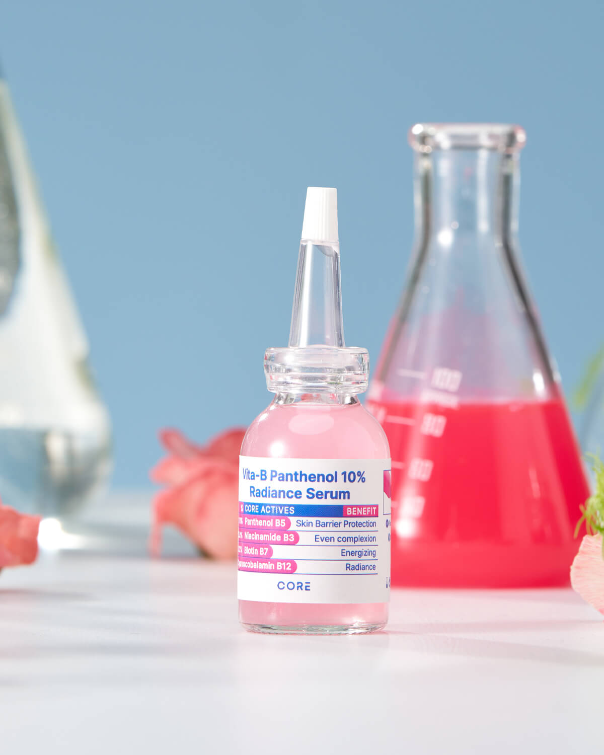 The pink serum bottle sits on a white surface, with a glass flask containing a bright pink liquid out of focus in the background, set against a blue wall.